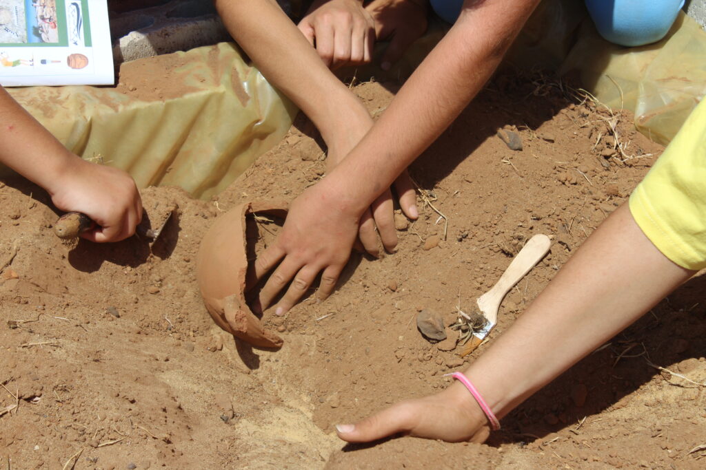 Four children's hands using archaeological tools to dig up and broken pot in a fake archaeological trench.