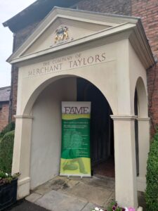 A white stone porch that says THE COMPANY OF MERCHANT TAYLORS, inside the porch is a banner that says FAME in red on a white background, with other text (un readable) in white on a green background below this.